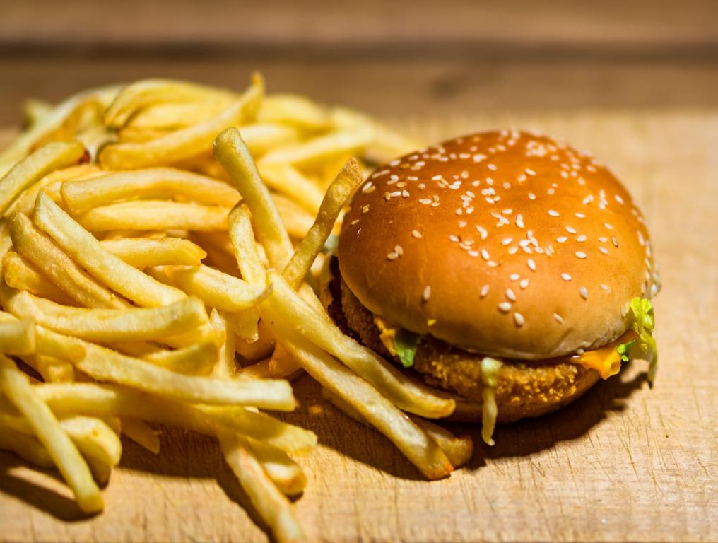 fRENCH FRIES SPILLED OUT ON A WOOD TABLE WITH A JUICY CHICKEN SANDWICH SITTING NEXT TO IT ON A SESAME SEED BUN.