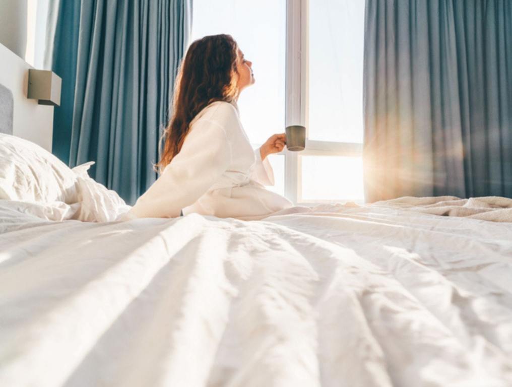 Pretty positive woman with long hair drinks water and sitting at bed in sunny lazy morning, affordable luxury hotel concept.